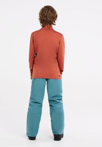 Langsleevige, roestkleurige top met een hoge hals die gecombineerd is met blauwe skibroeken. Het materiaal lijkt soepel te zijn, ontworpen voor actieve buitenactiviteiten.