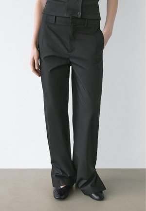 Donna che indossa pantaloni neri a gamba dritta a vita alta con tasche laterali e scarpe nere, in piedi davanti a uno sfondo bianco uniforme.