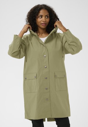 Egy nő beállítja az olíva zöld, hosszú esőkabátja kapucniját, amelynek elöl gombjai és nagy fedett zsebei vannak. Fekete nadrágot és fehér felsőt visel.