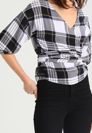 Mujer con blusa cruzada de cuadros en negro, blanco y gris, y pantalones negros, posando con una mano en la cadera sobre un fondo sencillo.