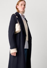 Saco trinchera navy con puños blancos, superpuesto a un suéter beige y una camisa de rayas azules. Sosteniendo un bolso color crema.