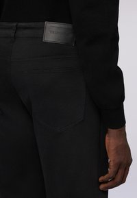 Jeans de mezclilla negra con un ajuste slim, que presentan costuras visibles, un bolsillo trasero y una etiqueta de marca de cuero texturizado en la cintura.