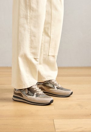 Baskets de couleur neutre avec des panneaux beige et marron portées avec un pantalon ample couleur crème clair, debout sur un sol en bois.