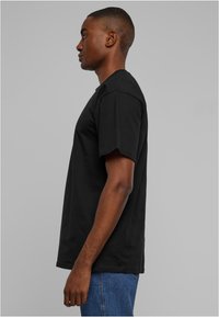 Tricou negru cu mânecă scurtă, cu guler rotund, fabricat din bumbac. Vedere laterală care arată o croială dreaptă și o potrivire lejeră, asortat cu blugi albaștri.