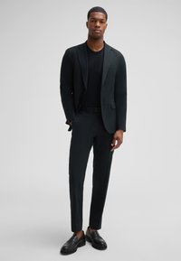 Schwarze Anzugjacke mit Reverskragen über einem schwarzen T-Shirt, kombiniert mit maßgeschneiderten schwarzen Hosen und schwarzen Loafers. Minimalistisches Design, glatte Textur.