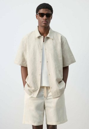 Joven con camisa beige de manga corta con estampado, pantalones cortos a juego, camiseta blanca y gafas de sol oscuras, de pie con las manos en los bolsillos.