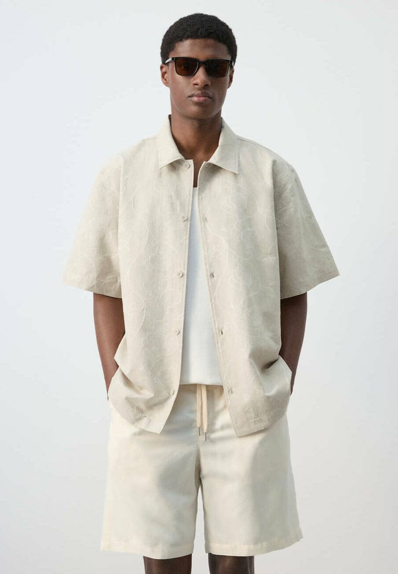 Jeune homme portant une chemise beige à manches courtes à motifs, un short assorti, un maillot blanc et des lunettes de soleil foncées, debout les mains dans les poches.