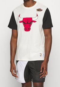 Bílé a černé tričko s krátkým rukávem s červeným logem Chicago Bulls na přední straně, vyrobené z bavlny, se zakulaceným výstřihem a znakem NBA.