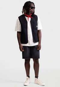 Colete preto sem mangas sobre uma t-shirt branca, combinado com calções de ganga escura e ténis brancos de cano alto. Um lenço vermelho na cabeça e óculos de sol completam o visual.