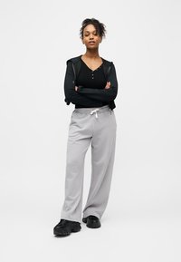 Jeune femme debout, les bras croisés, portant un sweat à capuche noir zippé, un haut noir, un pantalon de jogging gris et des baskets noires sur fond blanc uni.