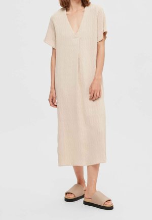 Kvinde iført en løstsiddende, beige, tekstureret midikjole med korte ærmer og V-hals, kombineret med beige plateausandaler på en hvid baggrund.