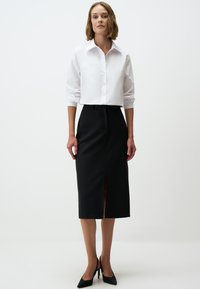 Weißes Langarmhemd mit Knöpfen, kombiniert mit einem schwarzen Midikleid mit Schlitz auf der Vorderseite, ergänzt durch schwarze spitze Absatzschuhe.