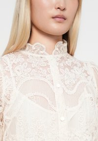 Blusa blanca de encaje con bordado floral, cuello alto y pequeños botones, fabricada con tejido transparente y delicados bordes festoneados.