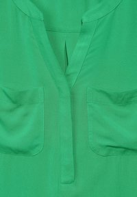 Vihreä pusero, jossa on V-mallinen kaula-aukko, kaksi etutaskua ja pehmeä, tekstuuri kangas. Sisältää pienen poimutuksen keskellä.