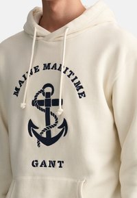 Kremowa bluza z kapturem wykonana z miękkiej tkaniny, z granatowym nadrukiem kotwicy z detalami liny oraz napisem "MAINE MARITIME GANT".