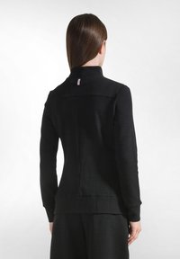 Giacca nera con un tessuto testurizzato, colletto alto e maniche lunghe. Presenta un piccolo etichetta sul collo posteriore e una silhouette aderente.
