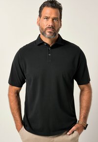Černé polo tričko s krátkým rukávem a límečkem na knoflíky, hladká textura a uvolněný střih. Nosené přes světle zbarvené kalhoty.