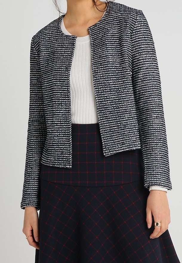 Femme portant une veste texturée rayée noire et blanche par-dessus un top côtelé blanc et une jupe noire avec un motif à grille rouge.
