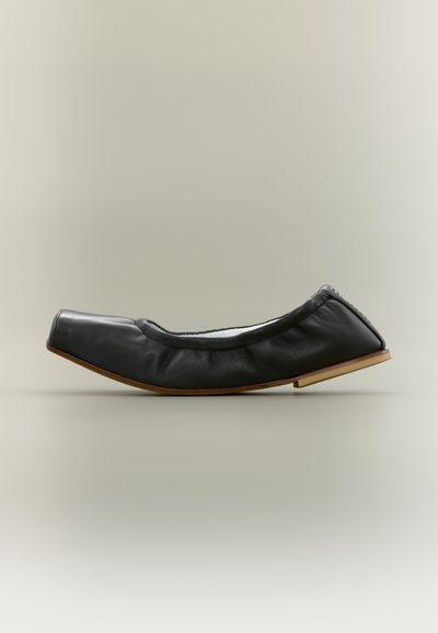 Chaussure plate en cuir noir souple avec bout carré et semelle en bois, conçue pour un confort facile à enfiler.