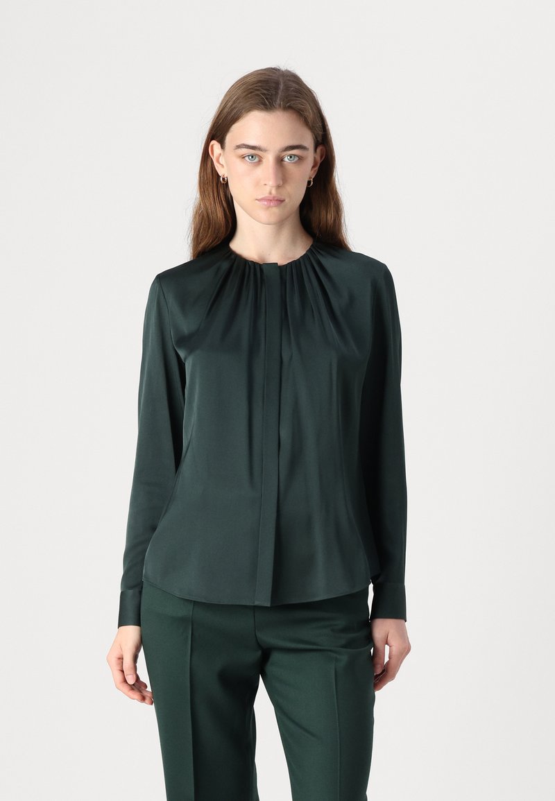 Mörkgrön silkblus med långa ärmar, med en plisserad halsringning och rakt snitt. Tillsammans med matchande byxor för en enhetlig look.