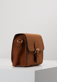 Brązowa zamszowa torba na ramię z klapą, złotymi okuciami i kontrastową czarną lamówką. Kompaktowy, kwadratowy kształt z ozdobną klamrą.