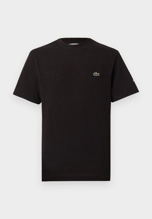 Due modelli indossano t-shirt e pantaloni neri Lacoste con sneakers bianche, posando su uno sfondo chiaro e uniforme.