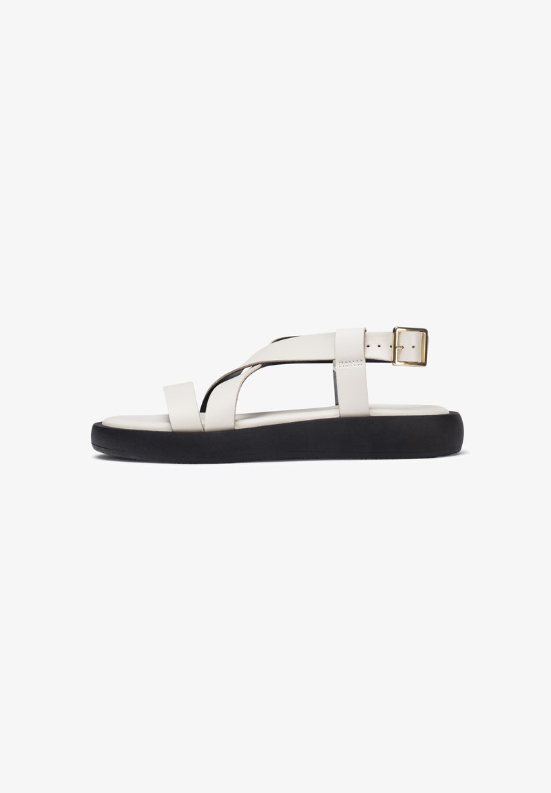 Sandal de cuero en tonos crema claros con correas cruzadas, hebilla ajustable en el tobillo y suela de goma negra. Diseño sencillo y plano con minimalismo en los detalles.