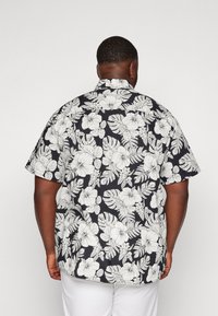 Camicia nera a maniche corte con un motivo floreale bianco, comprendente ibiscus e foglie, realizzata in tessuto leggero adatto per il clima caldo.
