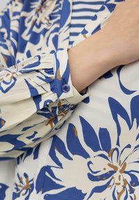 Chemise florale avec des motifs bleus, crèmes et bruns. Présente un poignet plissé avec un bouton bleu. Tissu lisse avec une forme semi-structurée.
