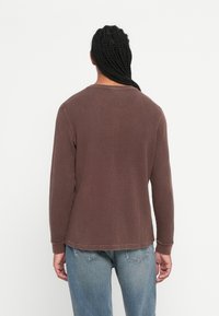 Person mit langen, geflochtenen Haaren, der ein braunes, langärmliges Waffelshirt und eine blaue Jeans trägt und mit dem Rücken zu einem weißen Hintergrund steht.