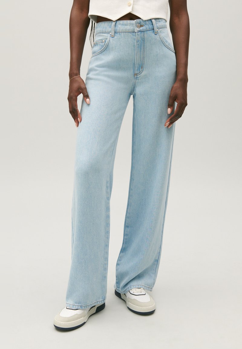 Personne portant un jeans large bleu clair et des baskets blanches, se tenant contre un fond clair uniforme.