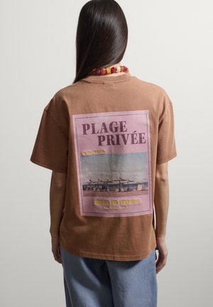 Persona con capelli lunghi e scuri che indossa una maglietta marrone con una scena di spiaggia e la scritta francese "Plage Privée" sulla schiena.