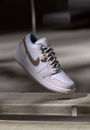 Baskets Nike blanches et beiges avec un charm en forme d'étoile sur les lacets, flottant au-dessus d'une surface en béton sur un fond flou.
