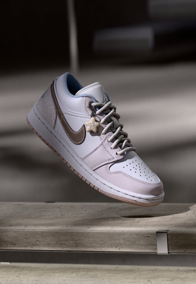 Baskets Nike blanches et beiges avec un charm en forme d'étoile sur les lacets, flottant au-dessus d'une surface en béton sur un fond flou.