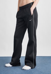 Pantaloni sportivi negri cu croi lejer, talie elastică, buzunare laterale și detalii albe pe cusături. Logo-ul mărcii este vizibil pe șold.