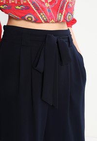 Mörkblå hög midja byxor med knytbart midjeband, plisserad front och sidofickor, tillverkade av ett lätt och slätt tyg.