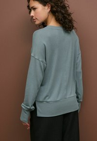 Světle tyrkysový oversized svetr s žebrovanými manžetami a lemem, hladká textura, design s pokleslými rameny, kombinovaný s černými kalhotami s pruhy.
