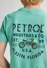 Tyrkysové bavlněné tričko s výraznou námořnickou grafikou, včetně nápisu "PETROL INDUSTRIES & CO." a ilustrace motocykla. Kulatý design písma.