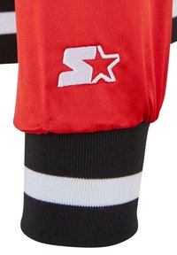 Rødt atletisk jakkeærme med sort og hvid stribet ribkant. Har hvidt broderet logo med en stjerne og bogstavet "S." Glat tekstur.