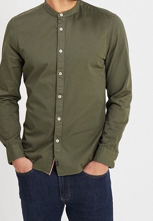 Homme portant une chemise boutonnée à col mandarin vert olive à manches longues et un jean bleu foncé, les mains dans les poches.