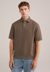 Camiseta polo de textura marrón con mangas cortas, tapeta de tres botones y cuello acanalado. Llevada sobre unos vaqueros azules claros.