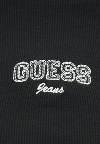 Tecido preto canelado com o texto bordado "GUESS" em letras brancas contornadas e "Jeans" em estilo cursivo branco menor.