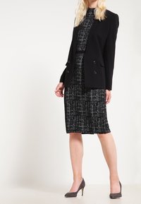 Skräddarsydd svart blazer med dubbelknäppning, bärs över en strukturerad svart-och-vit klänning, tillsammans med grå stilettklackar.