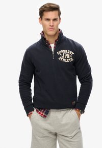 Suéter azul marino con cuello de cremallera, que presenta texto de logo color crema. Lleva encima una camisa a cuadros rojos. Unos pantalones de chándal grises completan el atuendo.