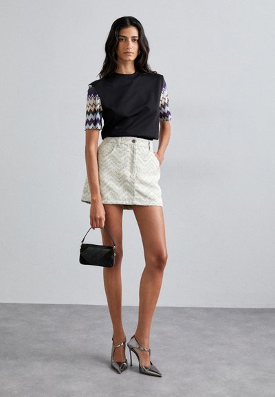 Haut noir avec des manches courtes à motif zigzag, mini-jupe texturée blanche, sac à main noir et chaussures à talons métalliques.