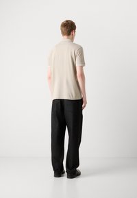 Beige Polohemd mit kontrastierenden Nähten, kombiniert mit lockeren schwarzen Hosen und schwarzen Schuhen, von hinten vor einem schlichten Hintergrund betrachtet.
