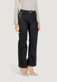 Nainen, jolla on ylhäältä korkeat mustat farkut koristeellisin nappilisäyksin lantiolla, beige ribattu neulepaita ja nude-huippukorkoiset kengät.