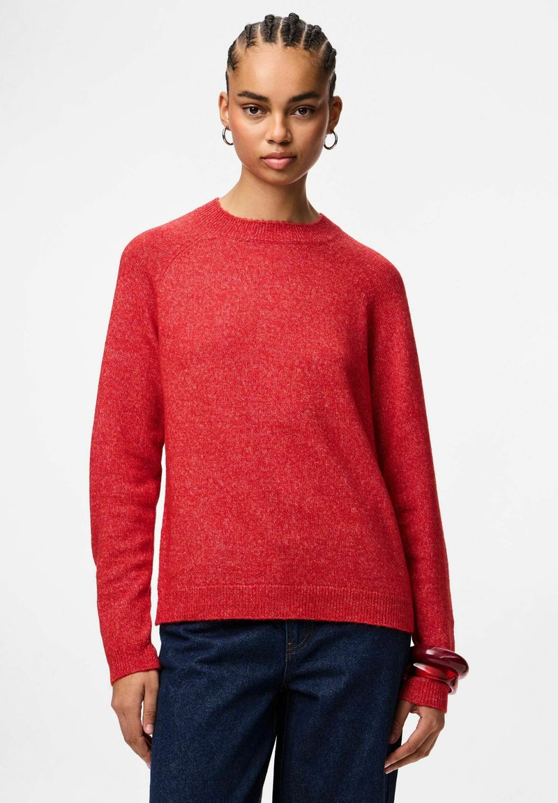 Raudonas megztinis su apykakle, ilgomis rankovėmis, braižytu kraštu ir subtilia tekstūra. Dėvimas su tamsiai mėlynomis džinsinėmis kelnėmis ir raudonu apyrankiu.