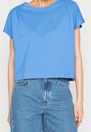 Persoon draagt een loszittend blauw cropped topje met korte mouwen, gecombineerd met een hoge taille blauwe spijkerbroek, zichtbaar van nek tot heupen.