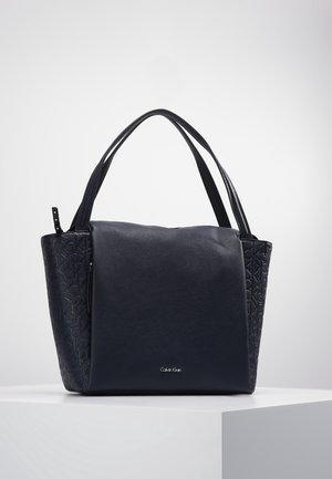 Sac à main en cuir noir Calvin Klein avec des côtés en logo embossé et des poignées doubles, posé sur un piédestal blanc contre un fond gris.